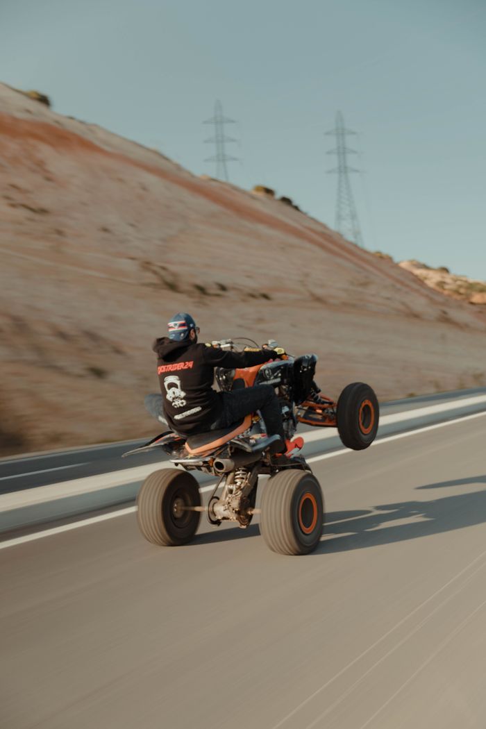 A man performing an impressive wheelie on an ATV on an open road against a desert backdrop.