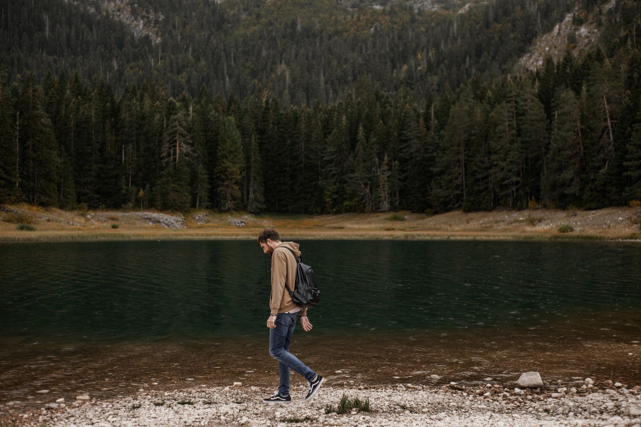 A lone man enjoys a walk by a tranquil mountain lake, surrounded by lush forest scenery.