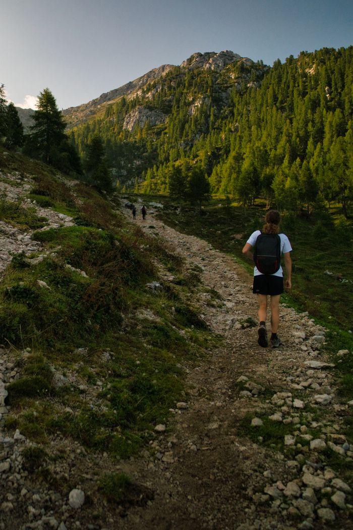 A person hiking along a rocky trail surrounded by lush green mountains under a blue sky.